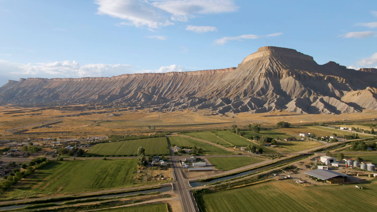 Opening frame over Palisades, Colorado showing green fields, a straight road, small farm buildings, tan mesa slopes, distant mountains, and no visible people.