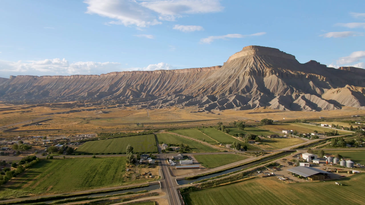 Wide aerial landscape with farmland, road, buildings, and tan mesa ridges.