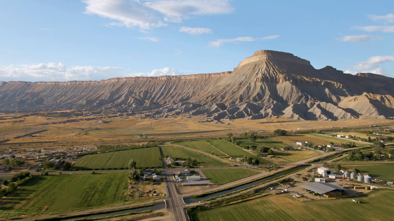 Third frame over Palisades, Colorado showing road lines crossing the foreground, rectangular green fields, small buildings, a broad tan ridge, and no visible people.