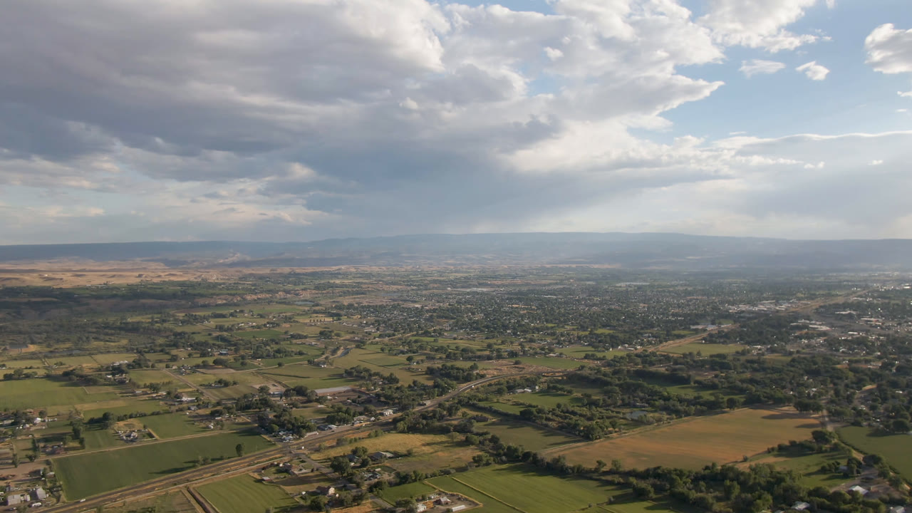 Opening aerial frame over Palisades, Colorado, with green and tan fields below, scattered roads and buildings in the distance, a broad cloudy sky above, and no visible people.