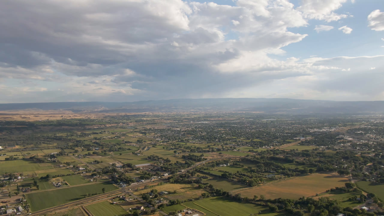 Aerial view across rural fields, roads, buildings, distant settlement, hills, and large clouds.