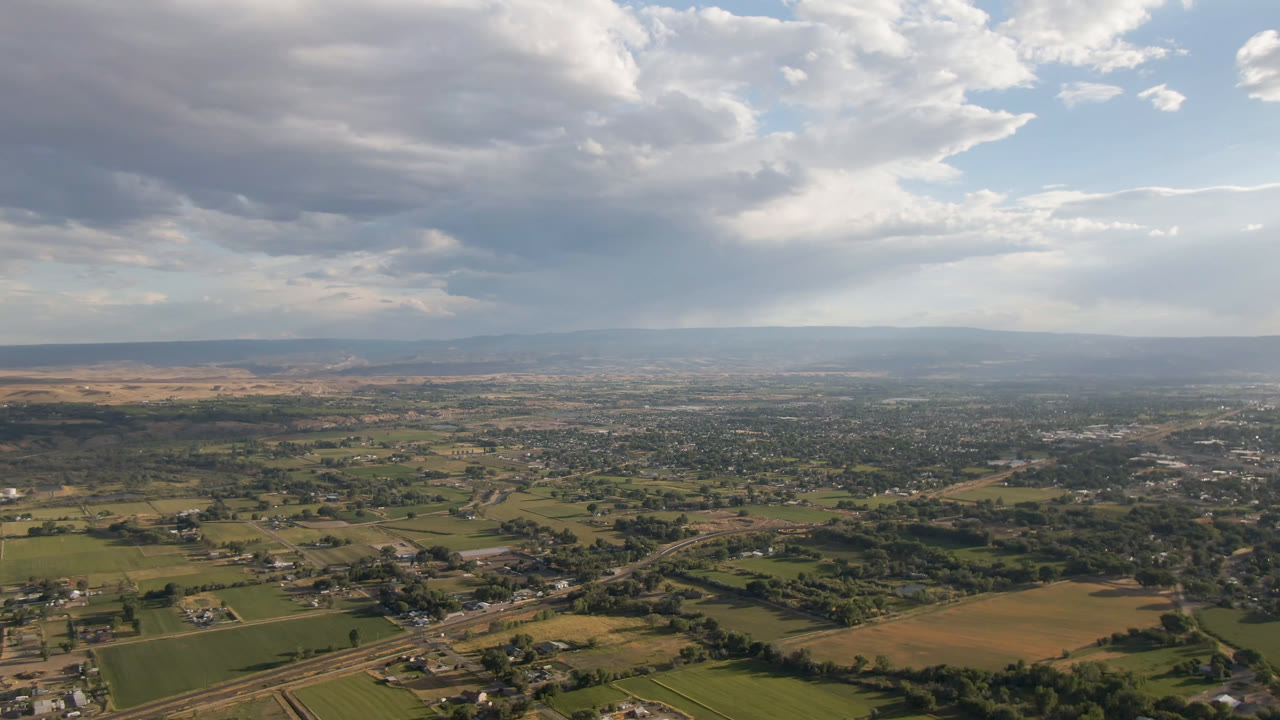 Mid-clip aerial frame over Palisades, Colorado, with road lines crossing fields in the foreground, scattered homes and town blocks beyond, distant hills, and no visible people.