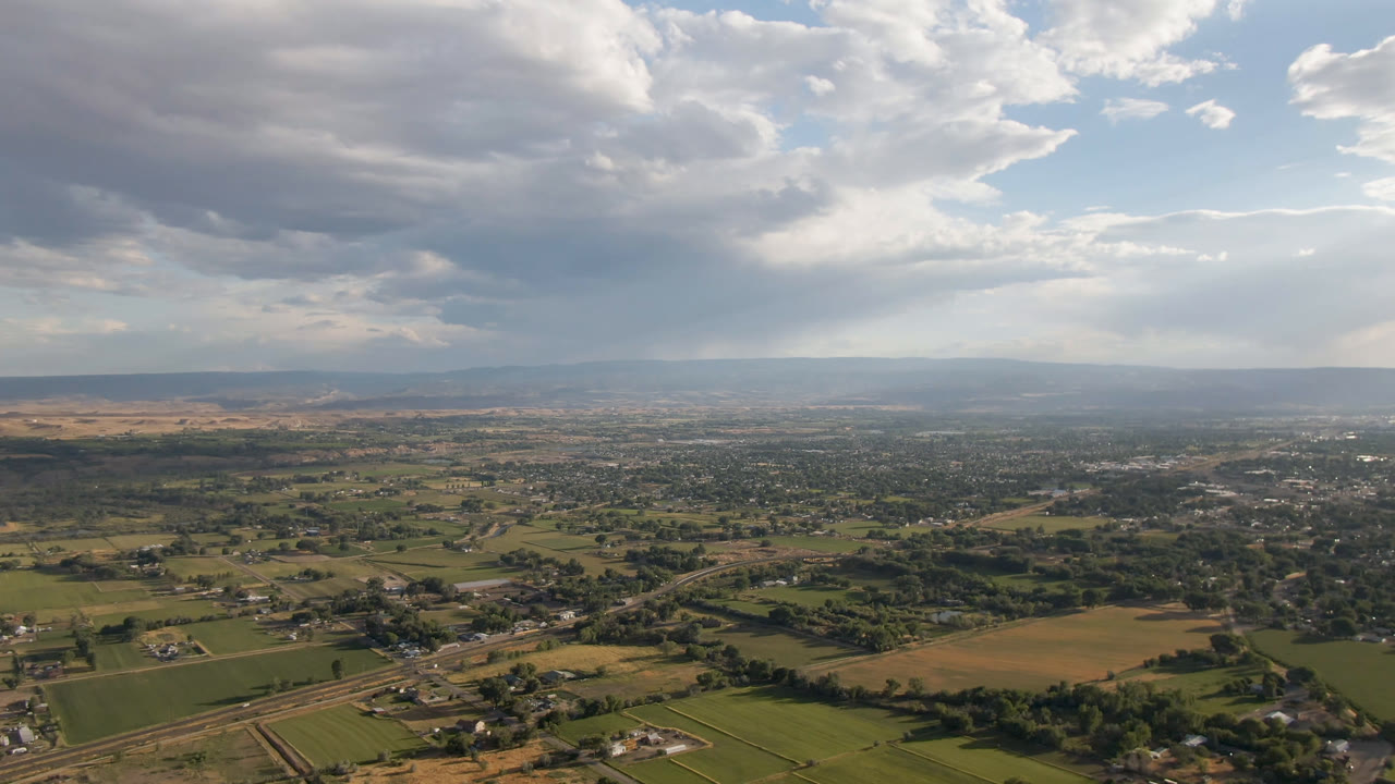 Wide aerial frame over Palisades, Colorado, with green fields, tan plots, scattered buildings, distant hills, and a large cloudy sky, with no visible people.