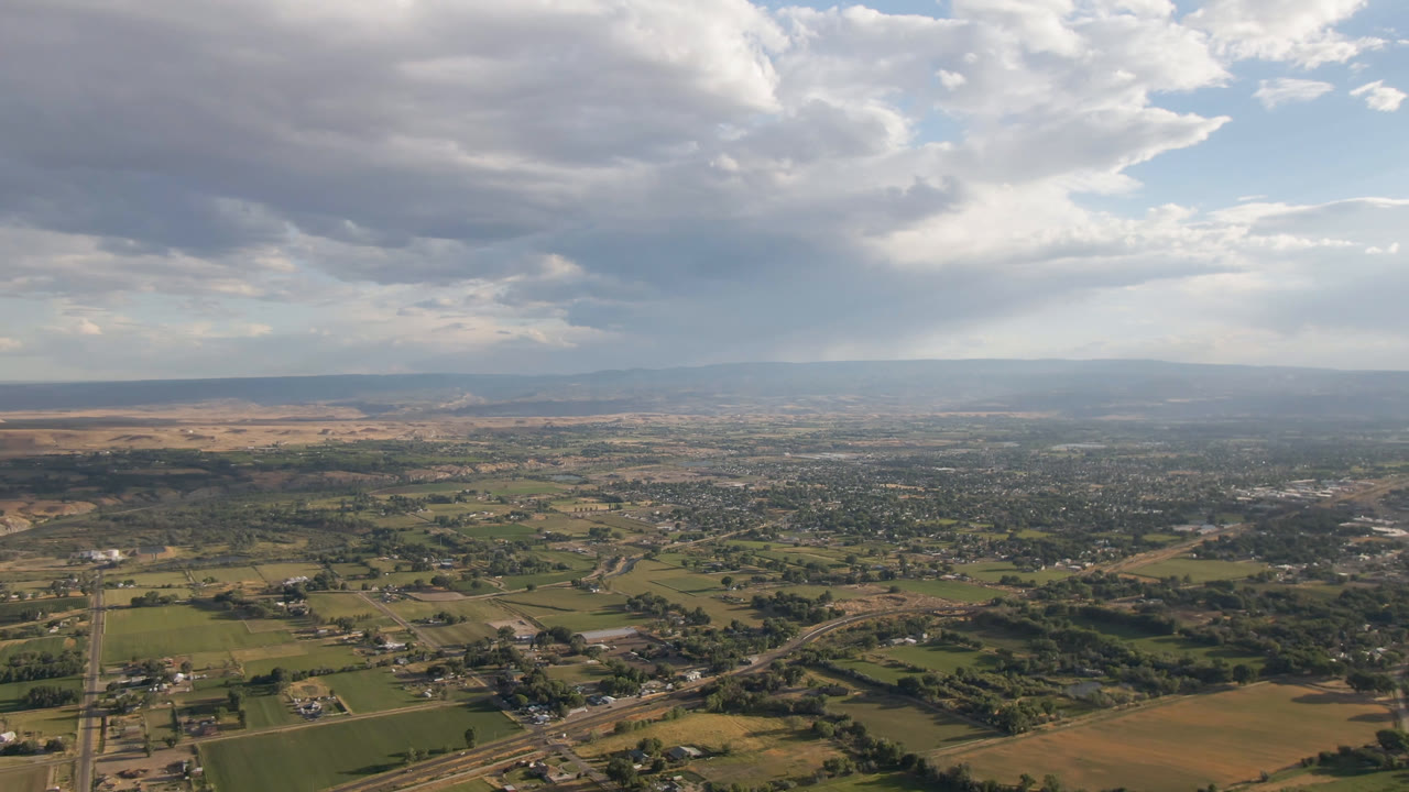 Wide aerial view over Palisades, Colorado, with green fields, tan farmland, roads, distant hills, and heavy gray clouds, with no visible people.