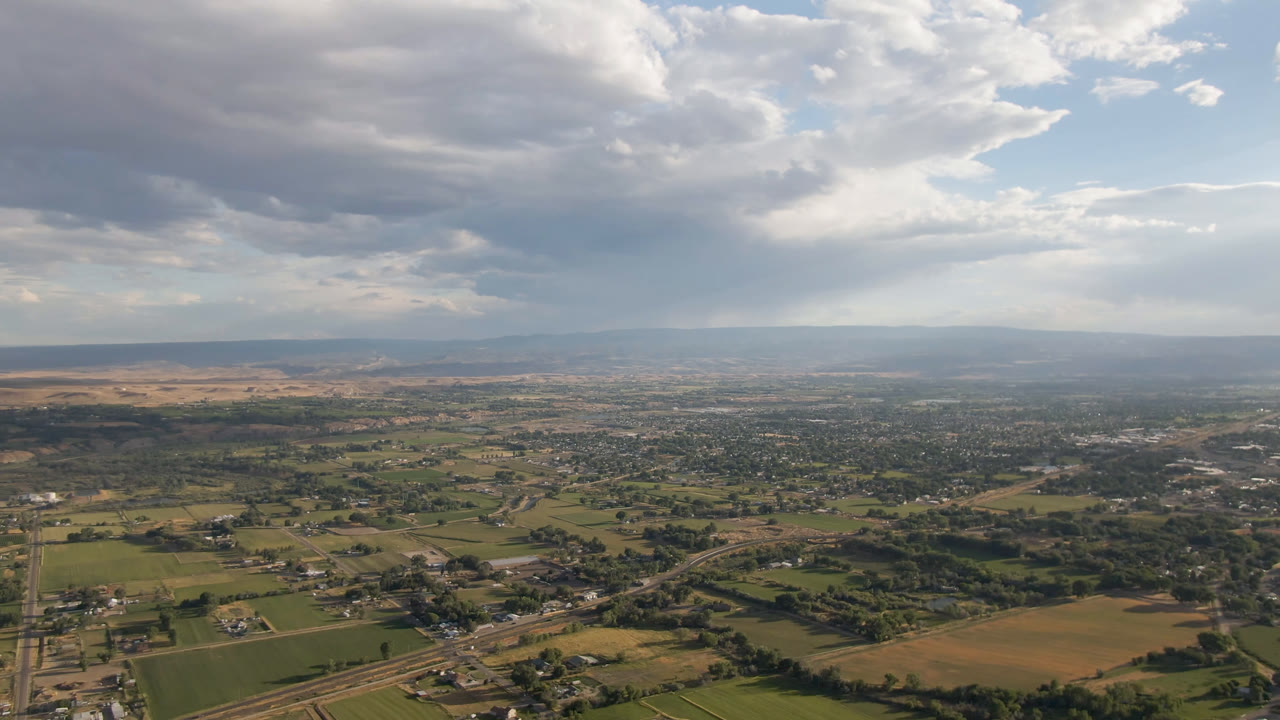 Opening aerial frame over Palisades, Colorado, showing green and tan fields, scattered buildings, roads, distant ridgelines, heavy clouds, and no visible people.