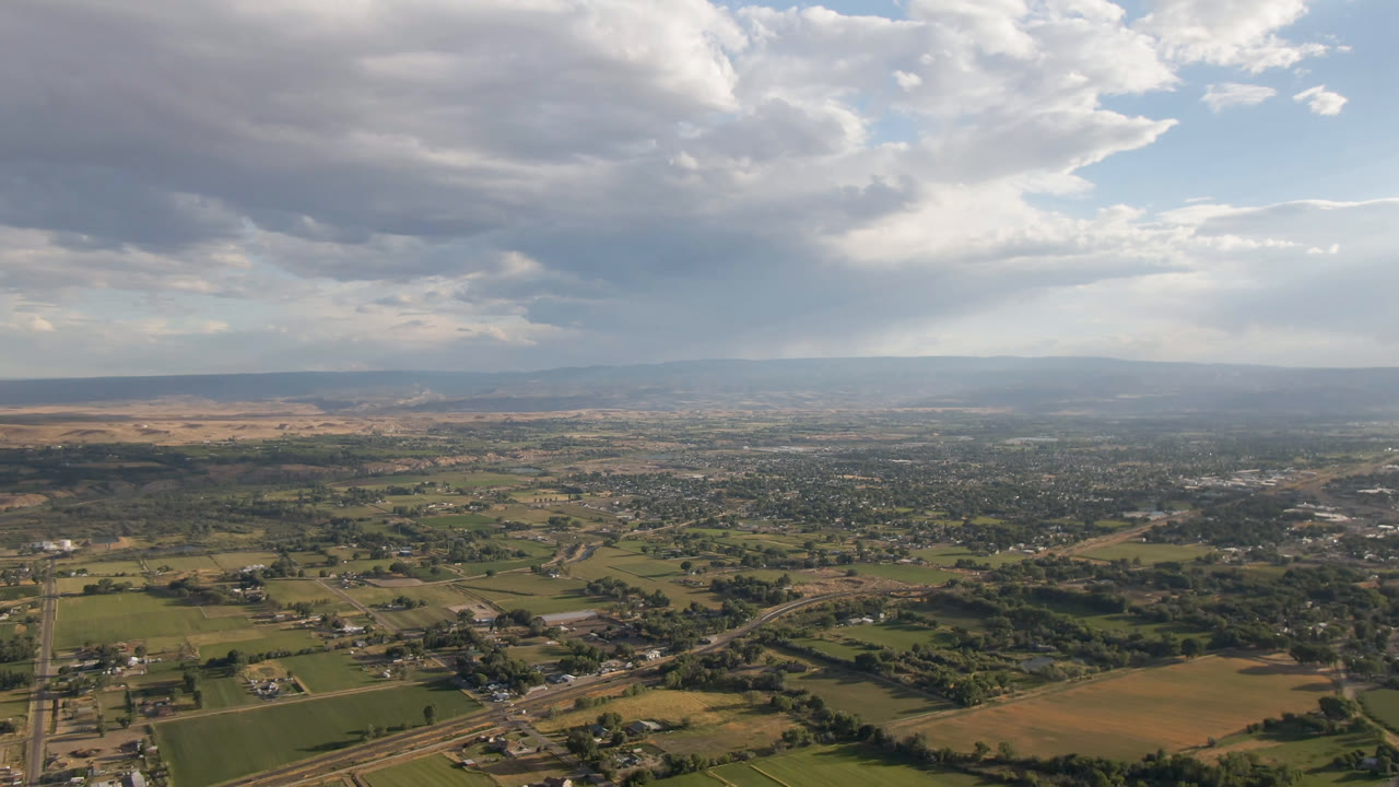 Second aerial frame over Palisades, Colorado, with more of the town grid visible across the midground, agricultural parcels below, distant hills, layered clouds, and no visible people.