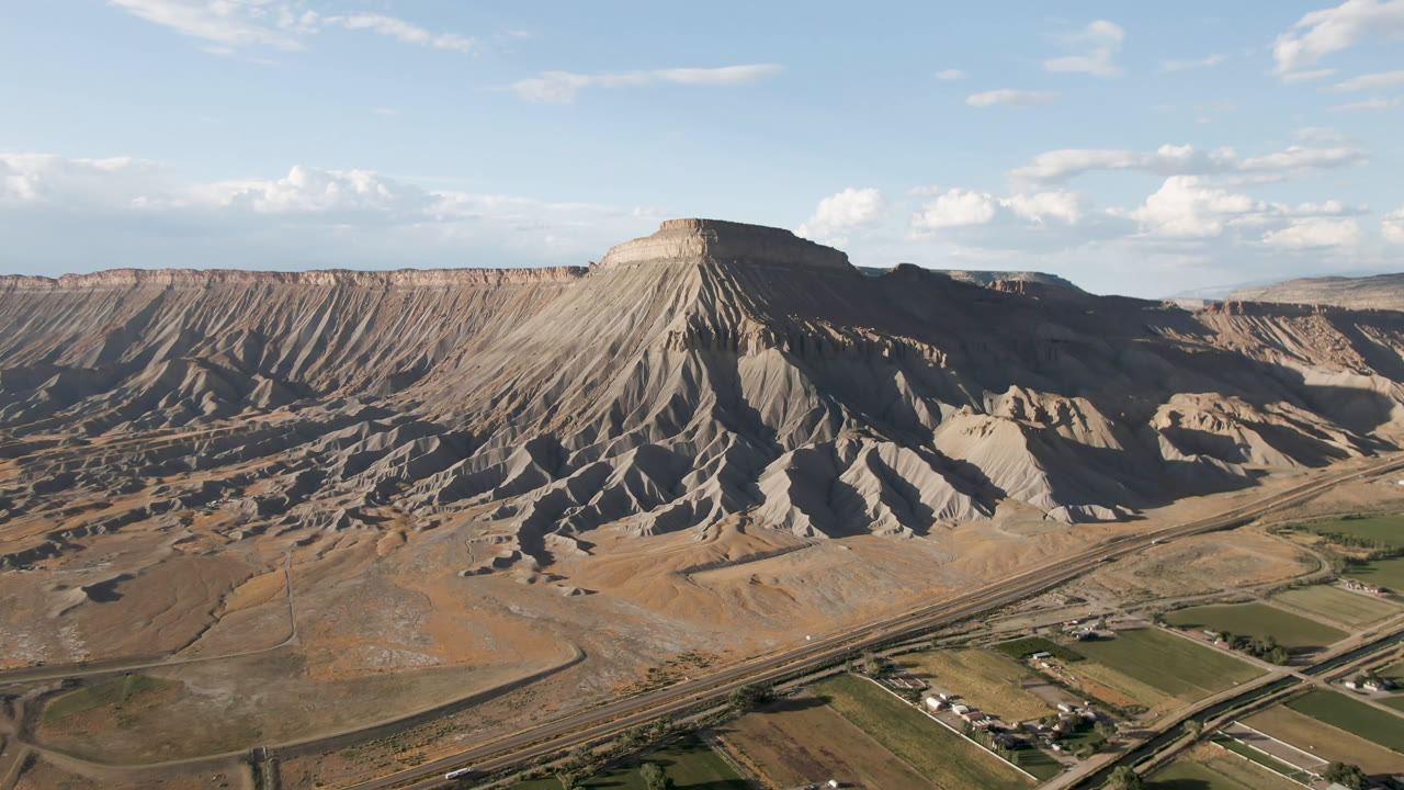 Aerial frame over Palisades, Colorado with the mesa centered, shadowed gullies on the slopes, a road network below, agricultural fields, scattered clouds, and no visible people.
