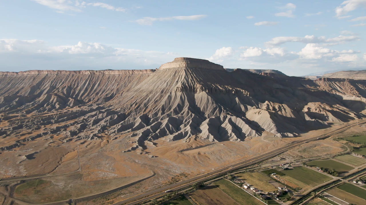 Second frame in Palisades, Colorado, with ridged gray-brown slopes, a diagonal road corridor, cultivated valley patches, distant mesas, and no visible people.
