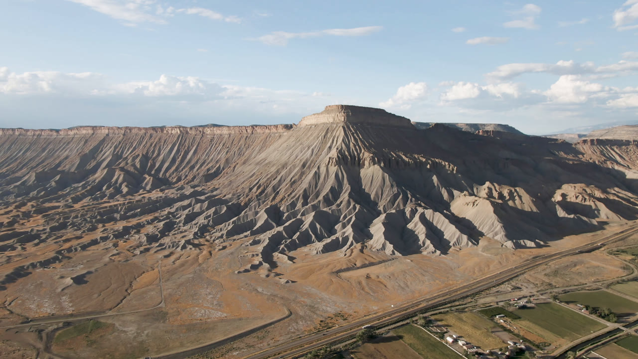 Wide aerial frame from Palisades, Colorado showing a flat-topped mesa, eroded slopes, valley roads, green fields, and no visible people.