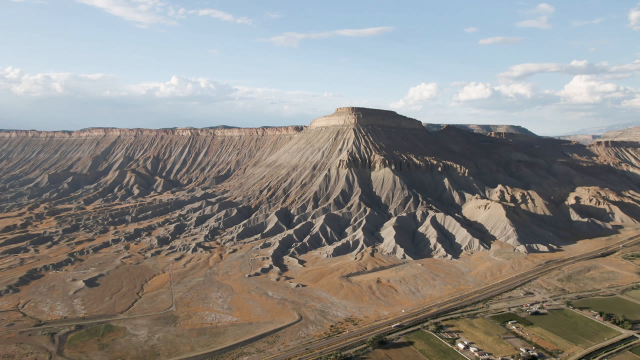 Aerial frame over Palisades, Colorado with a steep mesa face casting dark shadows, pale eroded gullies, valley roads, fields, and no visible people.