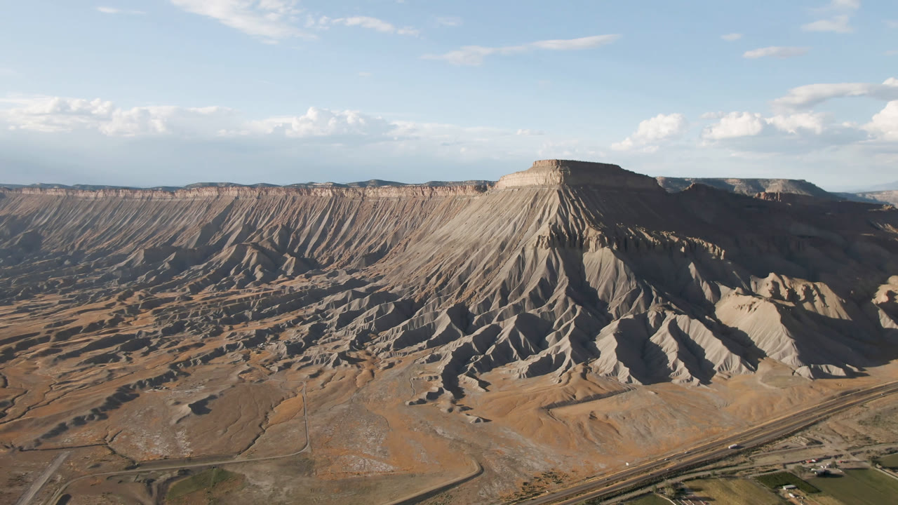 Aerial view of a steep gray and tan mesa slope in Palisades, Colorado, with sandy terrain below, scattered clouds above, and no visible people.