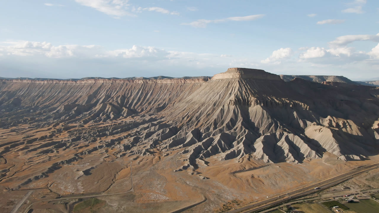Wide aerial frame in Palisades, Colorado, showing the eroded mesa wall stretching across the horizon, dry ground below, and no visible people.