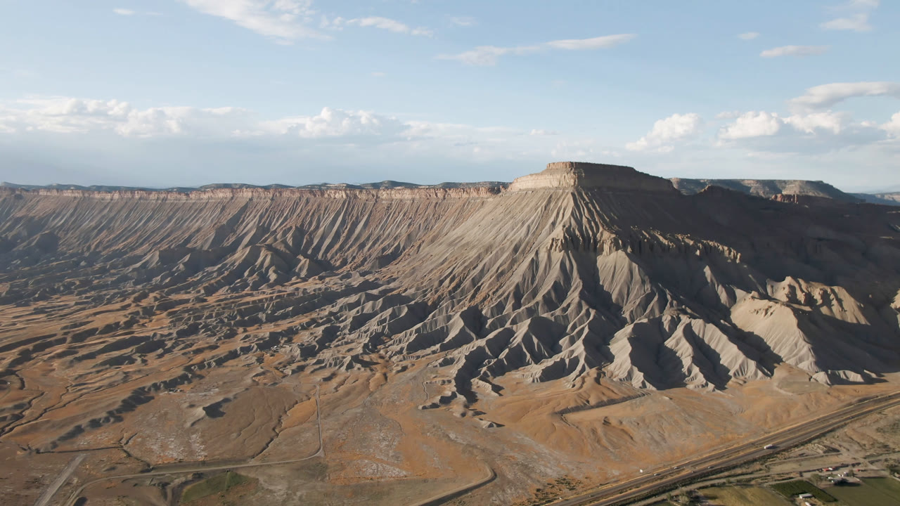 Aerial view in Palisades, Colorado, with a flat-topped mesa centered high in frame, road corridors at the base, shadowed gullies, and no visible people.