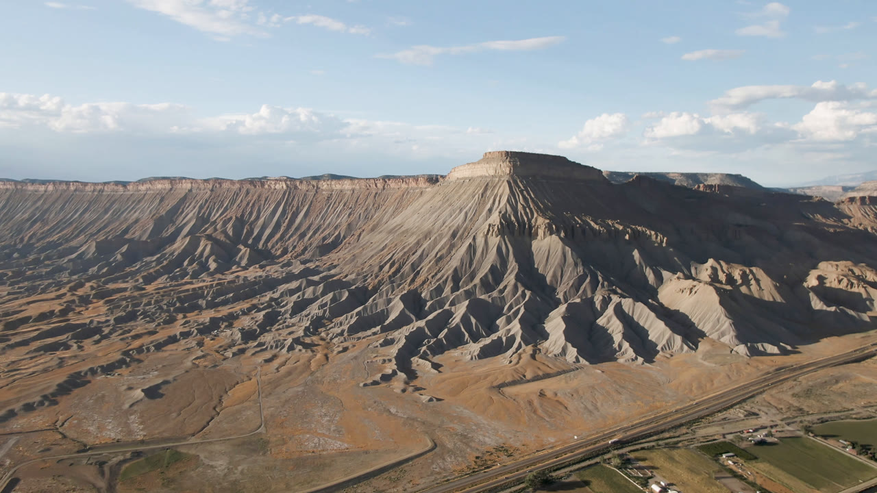 Opening frame of Palisades, Colorado from above, with a broad flat-topped mesa centered over gray-tan ridges, roads and small structures low in frame, and no visible people.