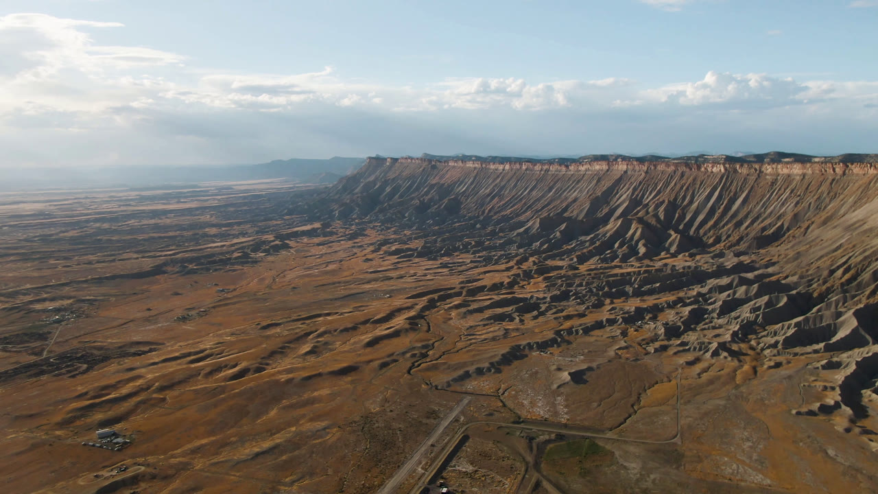 At 0:01, the Palisades, Colorado landscape remains wide, with the road network low in frame, a shadowed mesa face across the center, hazy flats beyond, and no visible people.