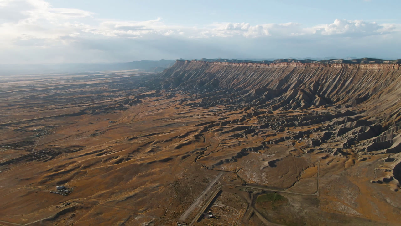 Opening aerial frame over Palisades, Colorado, showing tan mesa terrain, a long cliff band, winding roads, scattered structures, and no visible people.