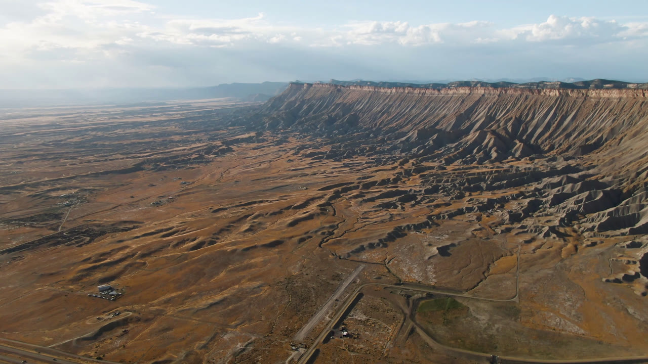 Aerial frame in Palisades, Colorado with a broad mesa wall across the horizon, eroded slopes below it, road lines in the foreground, and no visible people.
