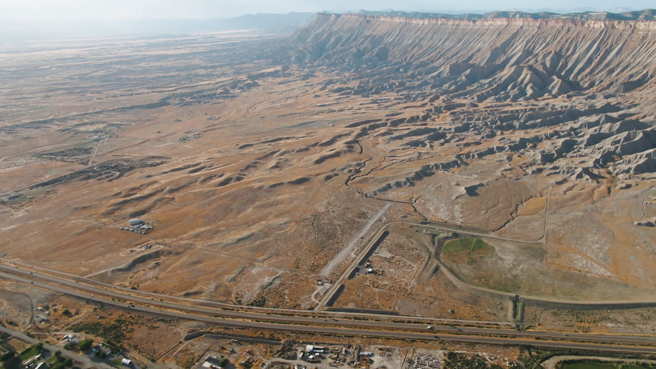 Opening aerial frame over Palisades, Colorado, showing highway lanes, a smaller road, tan open ground, green patches at the bottom edge, and a ridged mountain wall with no visible people.