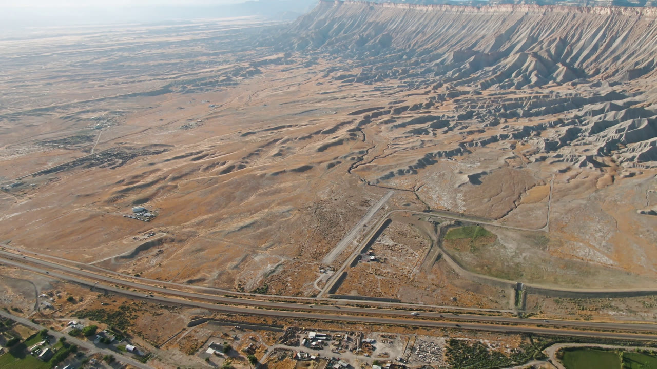 One second into the Palisades, Colorado aerial, the highway sits across the lower frame while pale sandy flats lead toward the eroded mountain ridge, with no visible people.