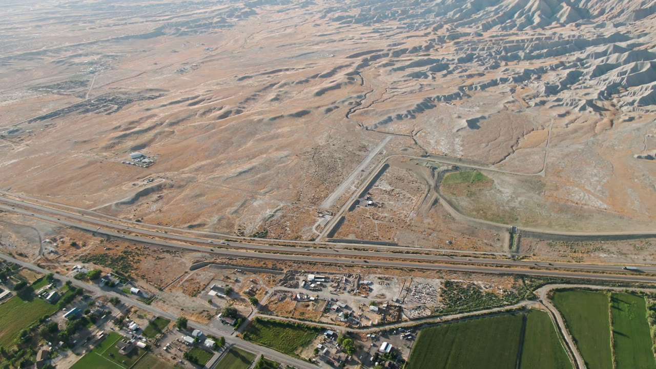 Aerial frame over Palisades, Colorado, with the highway running across the lower half, a diagonal road junction, green farm plots, rocky terrain, and no visible people.