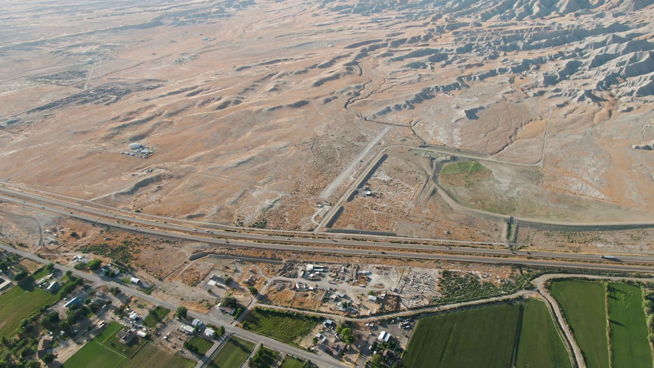 Overhead view in Palisades, Colorado, showing parallel highway lanes, a small cluster of buildings, green fields at the bottom edge, eroded hills beyond, and no visible people.