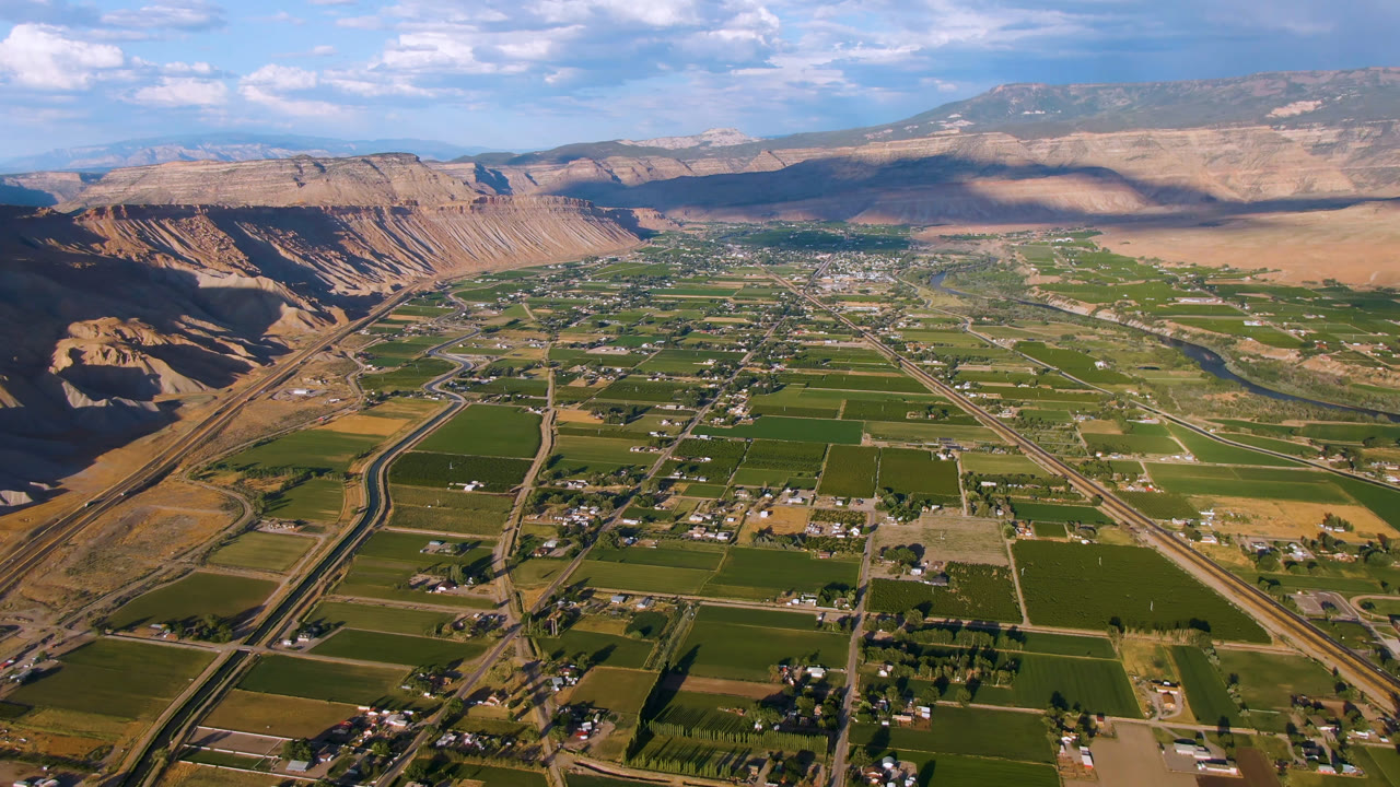 Opening aerial frame over Palisades, Colorado with green farm parcels, scattered buildings, winding roads, tan mesas, distant mountains, blue sky, and no visible people.