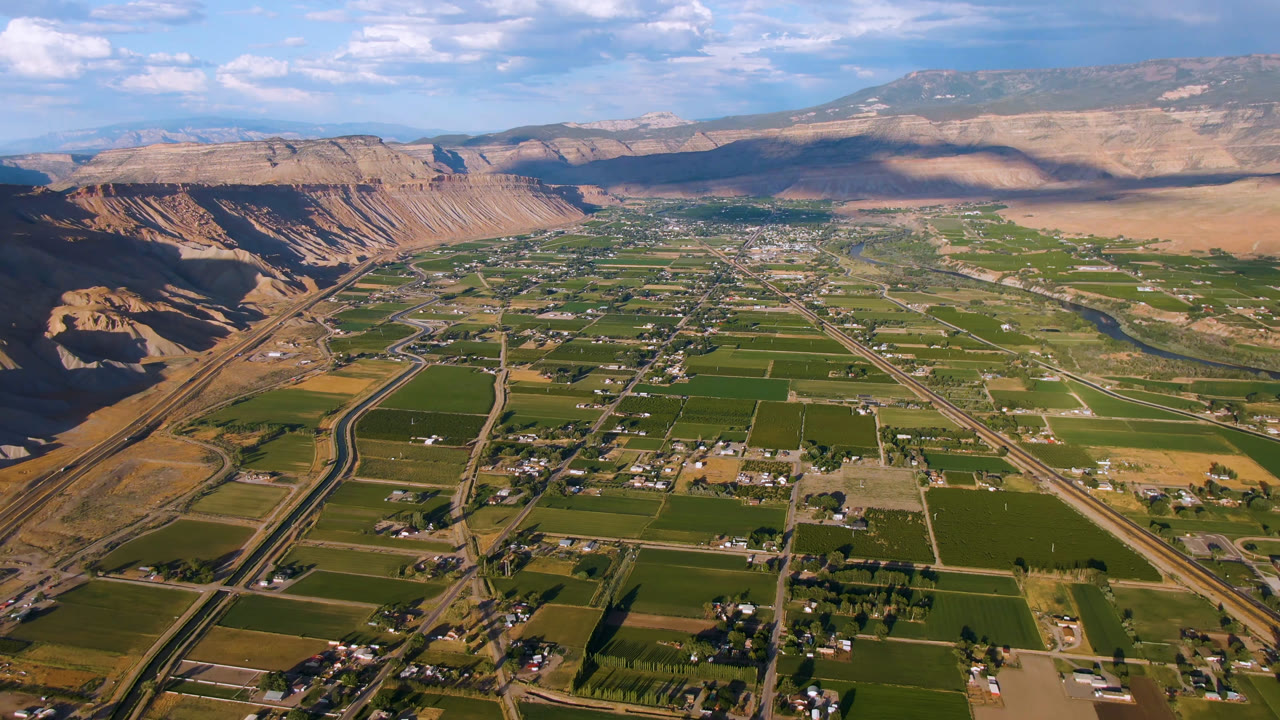 Aerial landscape with farmland blocks, scattered homes, roads, mesas, and clouds.