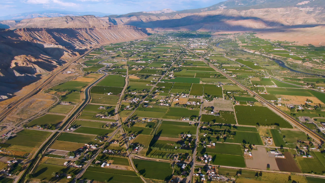 Opening aerial frame over Palisades, Colorado, with green farm blocks, small roads, scattered buildings, tan cliffs at left, distant mountains, and no visible people.