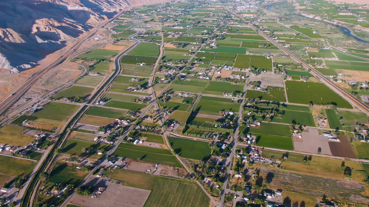 High aerial frame in Palisades, Colorado with a canal or divided road running through green fields, scattered buildings, dry hills, and no visible people.