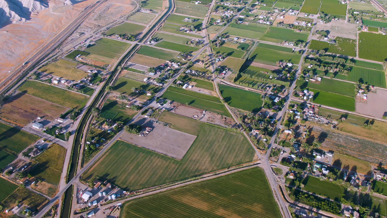 High aerial frame over Palisades, Colorado showing green fields, scattered homes, a road corridor, dry hills at the top edge, and no visible people.