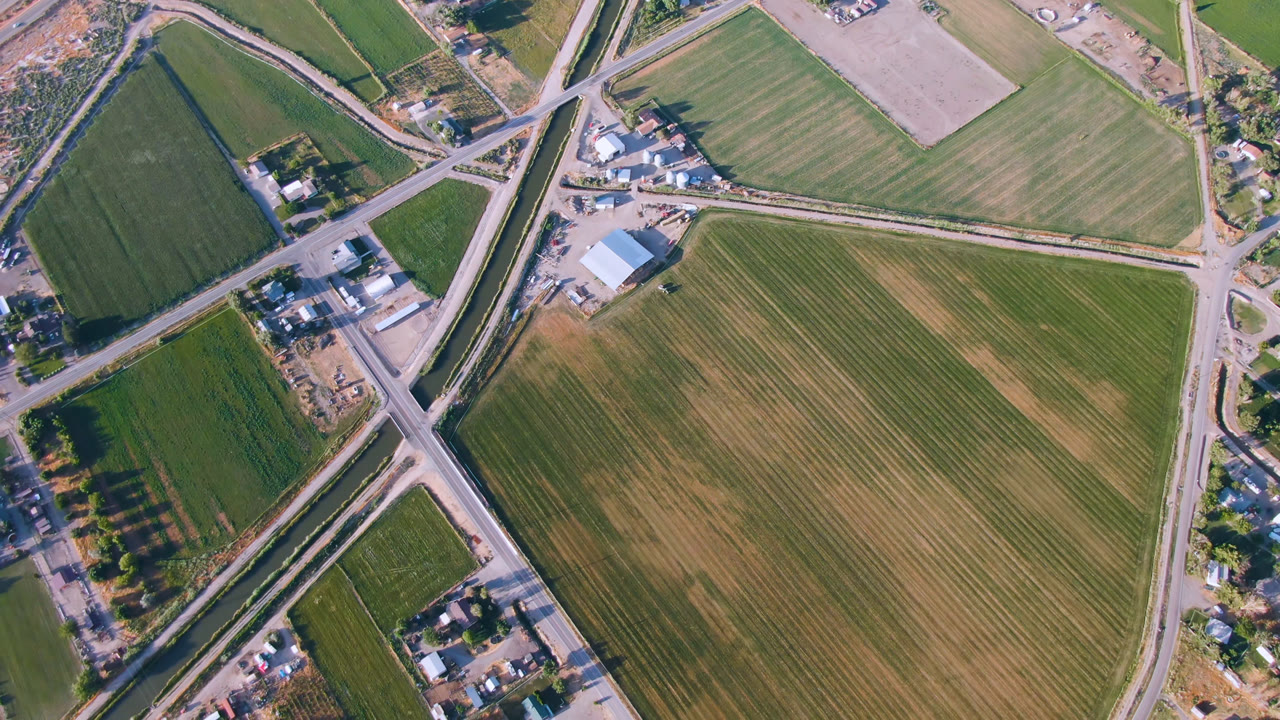 Aerial frame in Palisades, Colorado with a large tan field at right, green fields at left, a diagonal road junction, farm buildings, and no visible people.