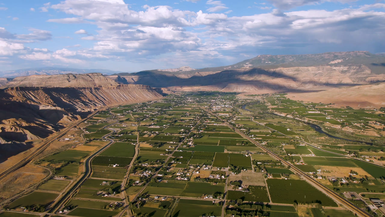 High aerial view over Palisades, Colorado, showing green agricultural fields, a road network, tan cliffs, distant mountains, and no visible people at the start of the clip.