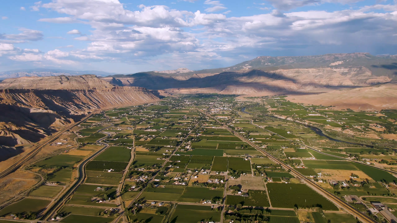 Wide aerial view in Palisades, Colorado, showing a winding road or canal corridor along the left valley edge, green fields across the floor, tan cliffs, and no visible people.