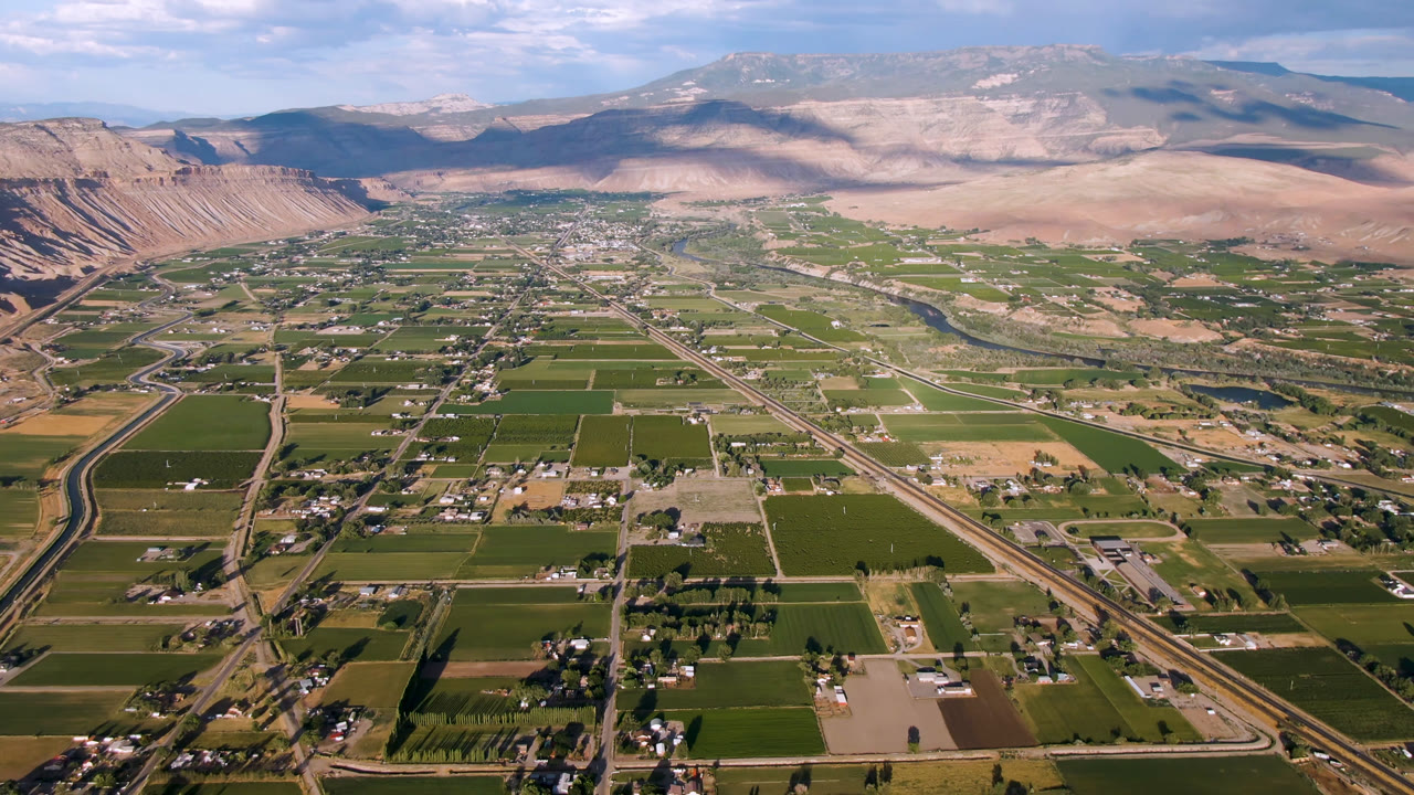 Aerial view of green fields, roads, scattered buildings, and mountains beyond a valley.