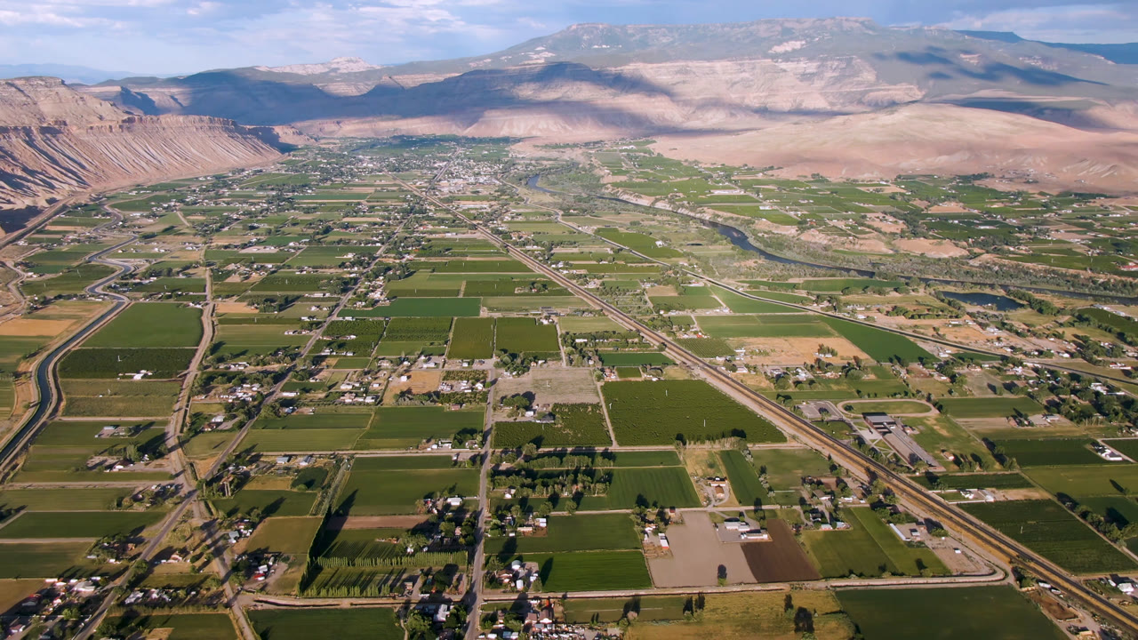 Aerial frame from Palisades, Colorado, with more of the central town area visible between green farm parcels, roads, mountain slopes, cloud shadows, and no visible people.