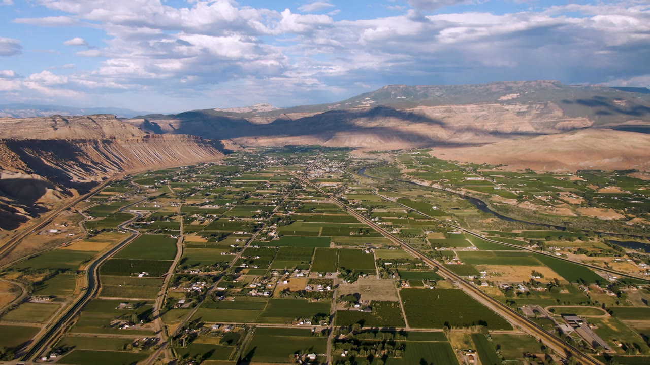 Wide aerial view of green fields, roads, ridges, and mountains under clouds.