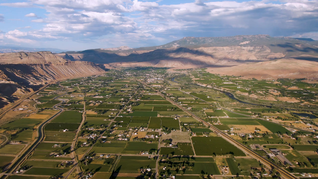 Aerial frame over Palisades, Colorado showing rectangular green fields, a diagonal road corridor, dry ridges at left, mountain horizon, cloud shadows, and no visible people.