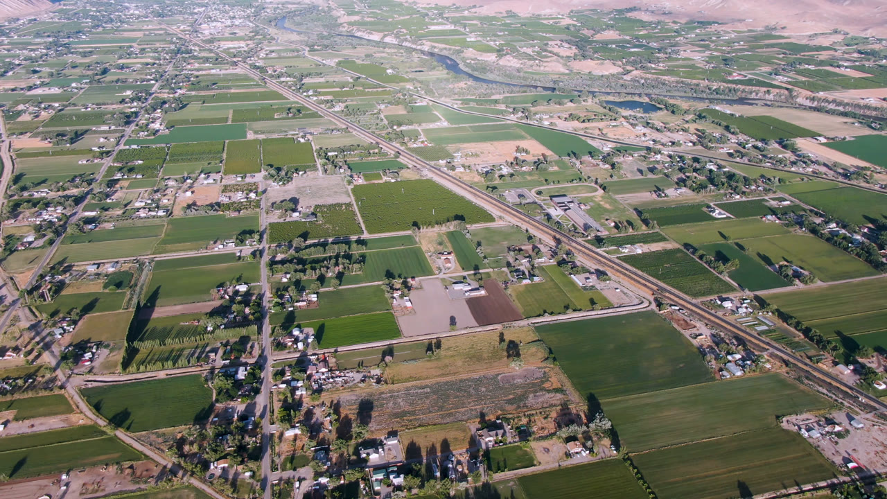 Opening aerial frame over Palisades, Colorado showing rectangular farm fields, diagonal road and rail corridors, a river near the top of frame, distant hills, and no visible people.