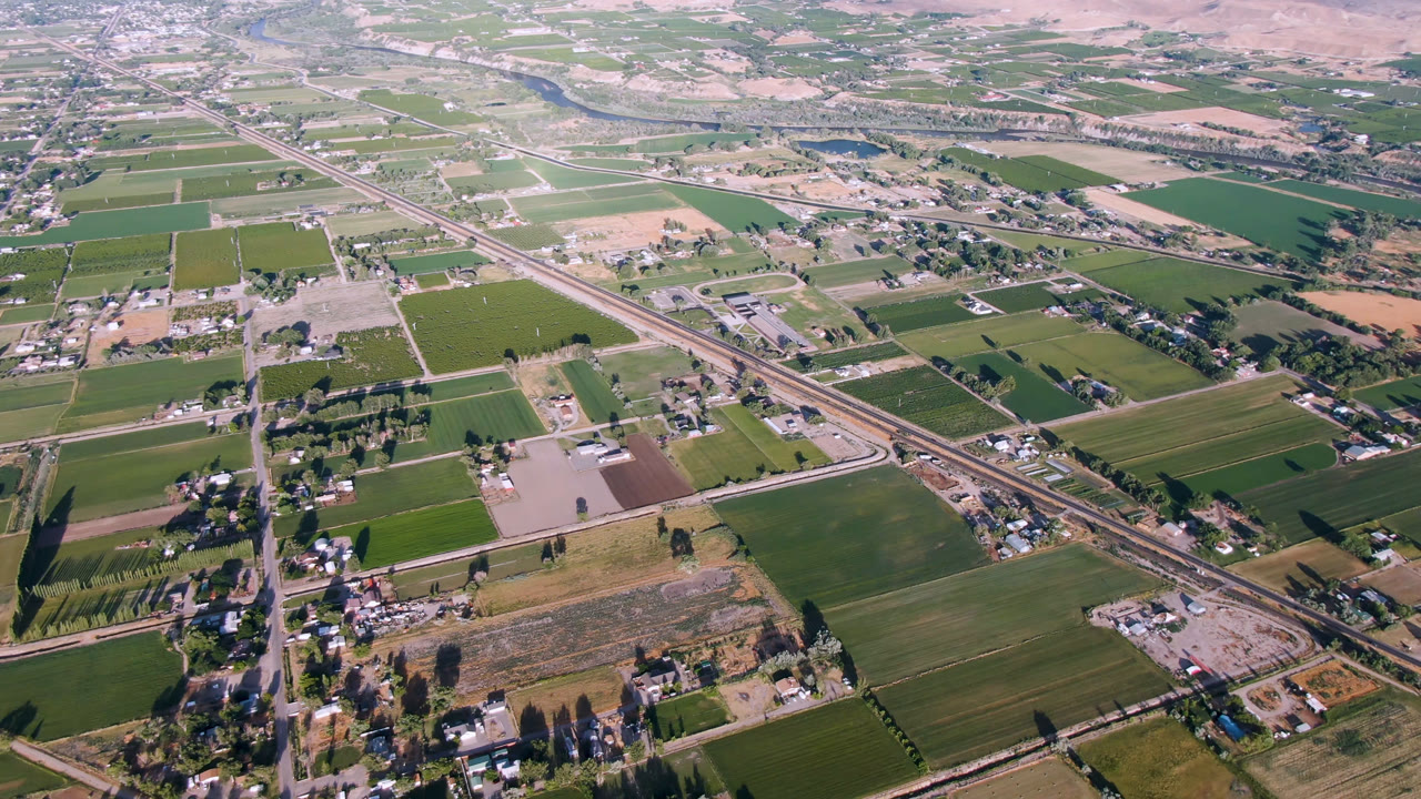 Second aerial frame over Palisades, Colorado, showing patchwork fields, a diagonal road or rail corridor, clustered rural buildings, dry background hills, and no visible people.
