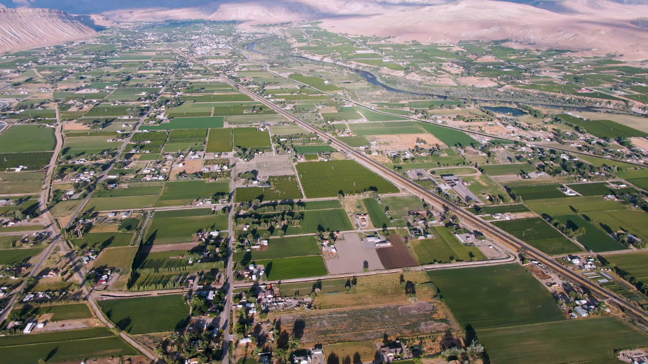 High aerial frame over Palisades, Colorado, showing rectangular green fields, tan parcels, a diagonal road line, small building clusters, distant mountains, and no visible people.