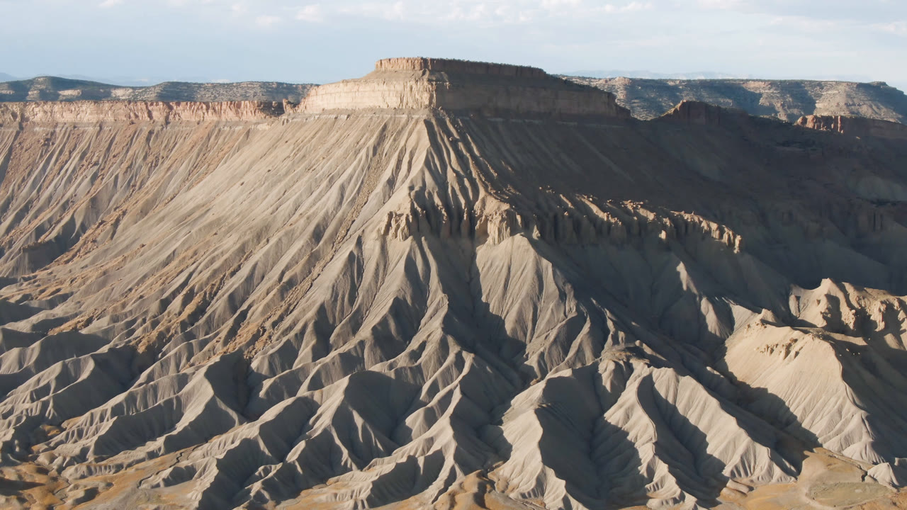 Wide aerial view of an eroded mesa face in Palisades, Colorado, with tan ridges, shadowed gullies, distant plateaus, blue sky, and no visible people.