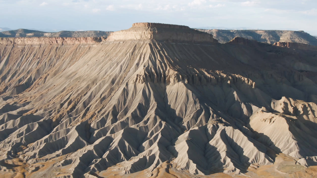 Aerial frame over Palisades, Colorado showing the mesa's vertical erosion ribs, gray-tan slopes, dry basin floor, distant ridgelines, and no visible people.