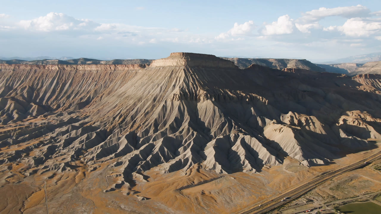 Wide aerial frame over Palisades, Colorado showing a flat-topped mesa, deeply eroded slopes, valley roads, scattered clouds, and no visible people.