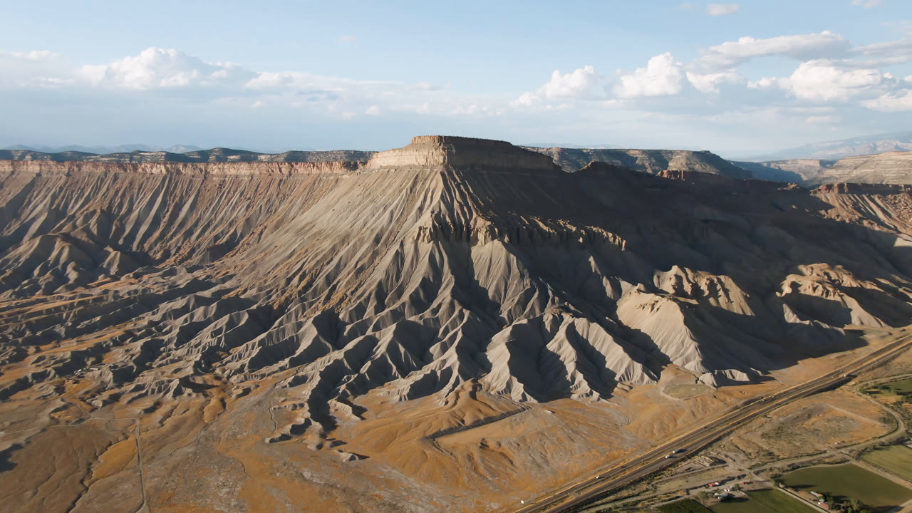Aerial view in Palisades, Colorado with the mesa centered, shadowed gullies dropping toward a road corridor, distant plateaus, and no visible people.