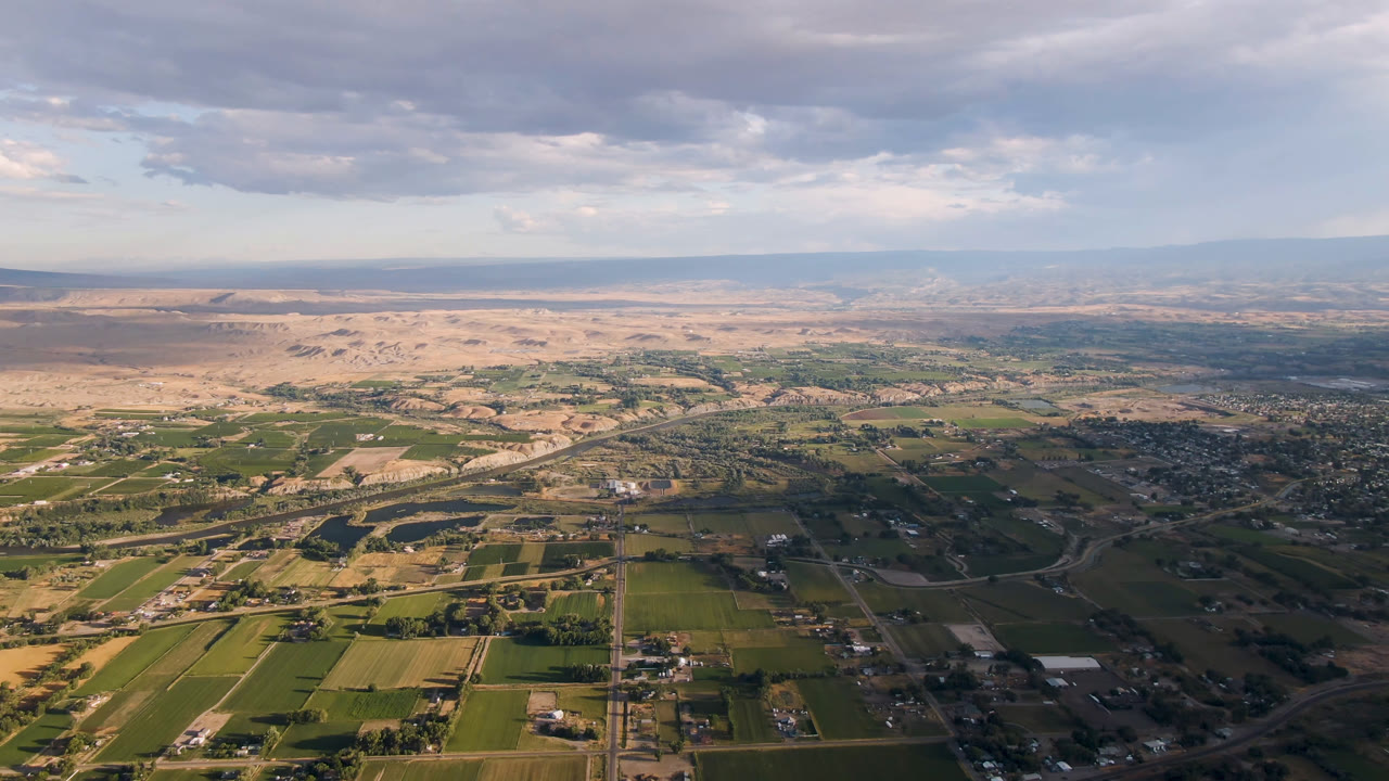 Wide aerial frame over Palisades, Colorado with green fields in front, roads and scattered buildings across the valley, pale desert terrain beyond, and no visible people.