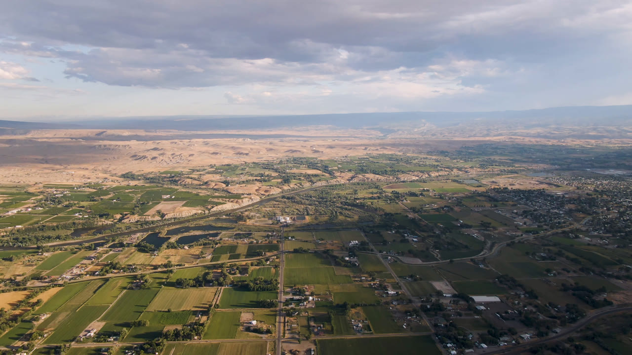 Aerial view of Palisades, Colorado showing patchwork agricultural fields, a road corridor, clustered buildings to the right, dry hills in the distance, and no visible people.
