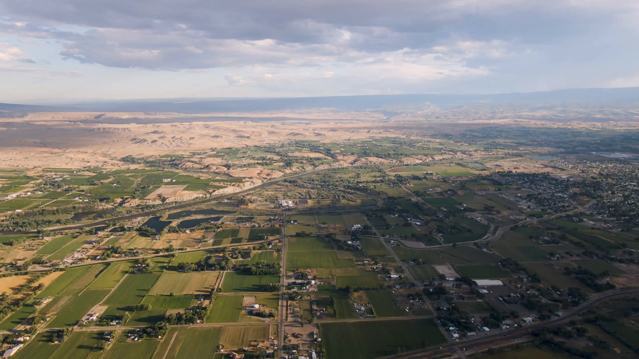 Aerial view of farm fields, roads, river bends, desert flats, mountains, and clouded sky.