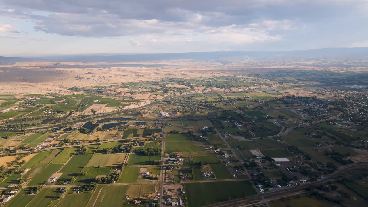 Aerial frame at 0:01 over Palisades, Colorado, showing a wider sweep of rectangular fields, small structures, intersecting roads, river curves, pale desert terrain, distant mountains, and no visible people.