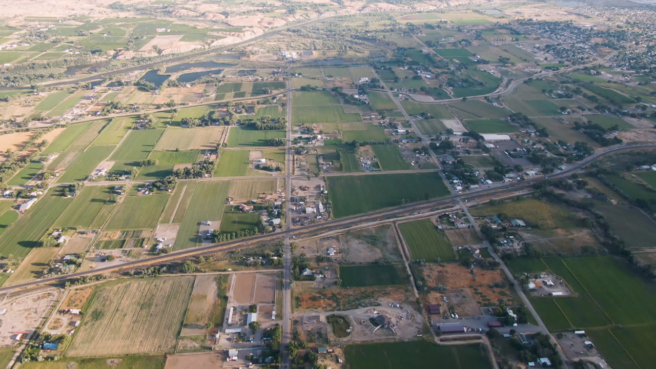 Second aerial frame of Palisades, Colorado with green and tan fields, small structures, straight roads, the diagonal rail or roadway line, distant settlement, and no visible people.