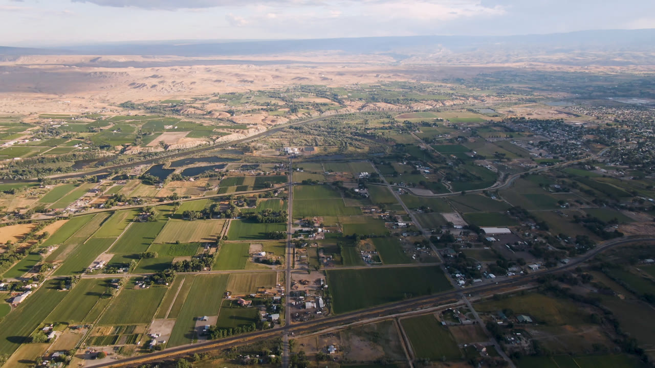 Wide aerial frame over Palisades, Colorado, showing green farm plots, scattered rural buildings, a diagonal highway, desert hills in the distance, and no visible people.